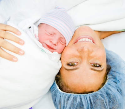 Image shows a woman lying down, post caesarean birth, smiling at the camera whilst holding her swaddled new baby next to her head.